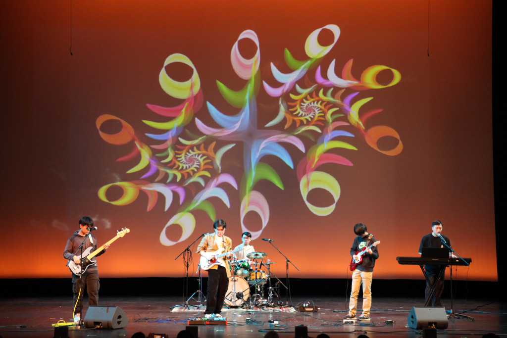 A rock band performs on stage while colorful rainbow swirls fill a screen behind them.