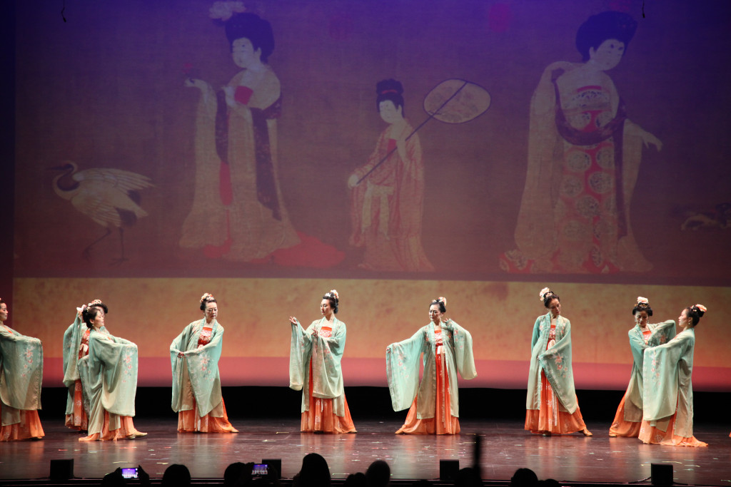 Seven women wearing traditional Chinese dress hold poses on a stage. Behind them is a projection of a classical Chinese painting showing women dressed and posed in the same style.