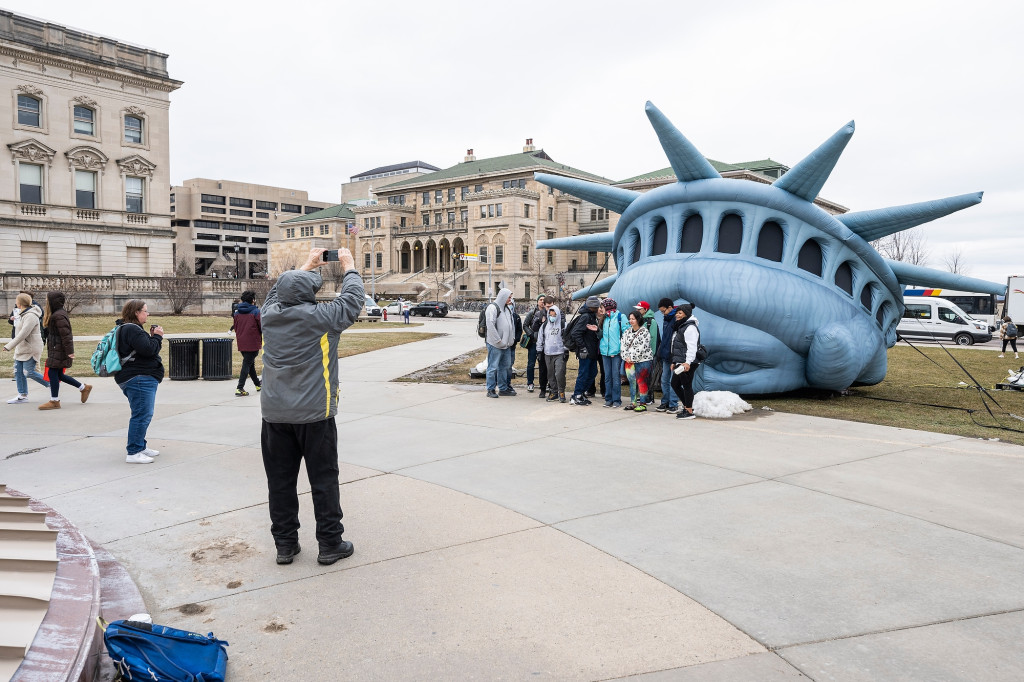 A person takes a photo of a group of people standing in front of an inflatable replica of the Statue of Liberty's head.