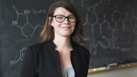 A photo portrait of Amy Weeks smiling. Behind her is a black chalkboard with drawings of chemical bonds.