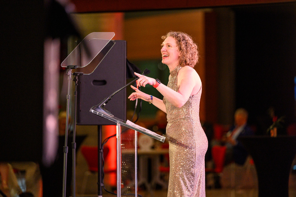Photo of Steffi Diem wearing a formal sequins gown and speaking to a large crowd from a podium.