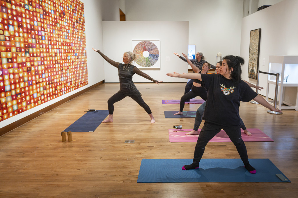 An instructor leads a small yoga class in warrior one pose as they stand in an art gallery surrounded by colorful paintings.