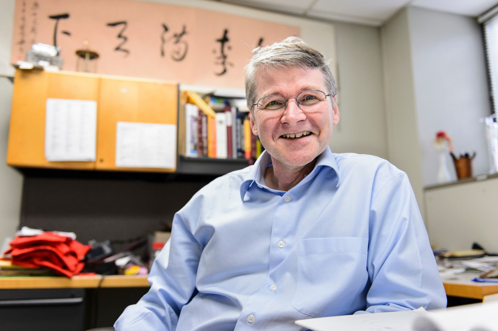 A man wearing a blue shirt is sitting in an office, in front of a bookshelf.