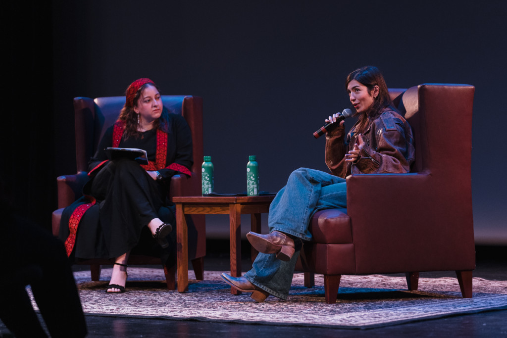 Two women sit on stage in chairs. One holds a microphone as part of a group discussion.