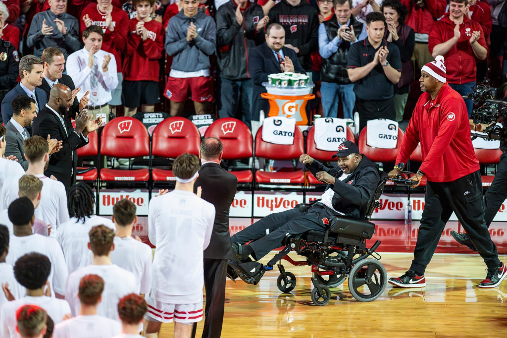 A group of players stand and applaud a man in a wheelchair on a basketball court.