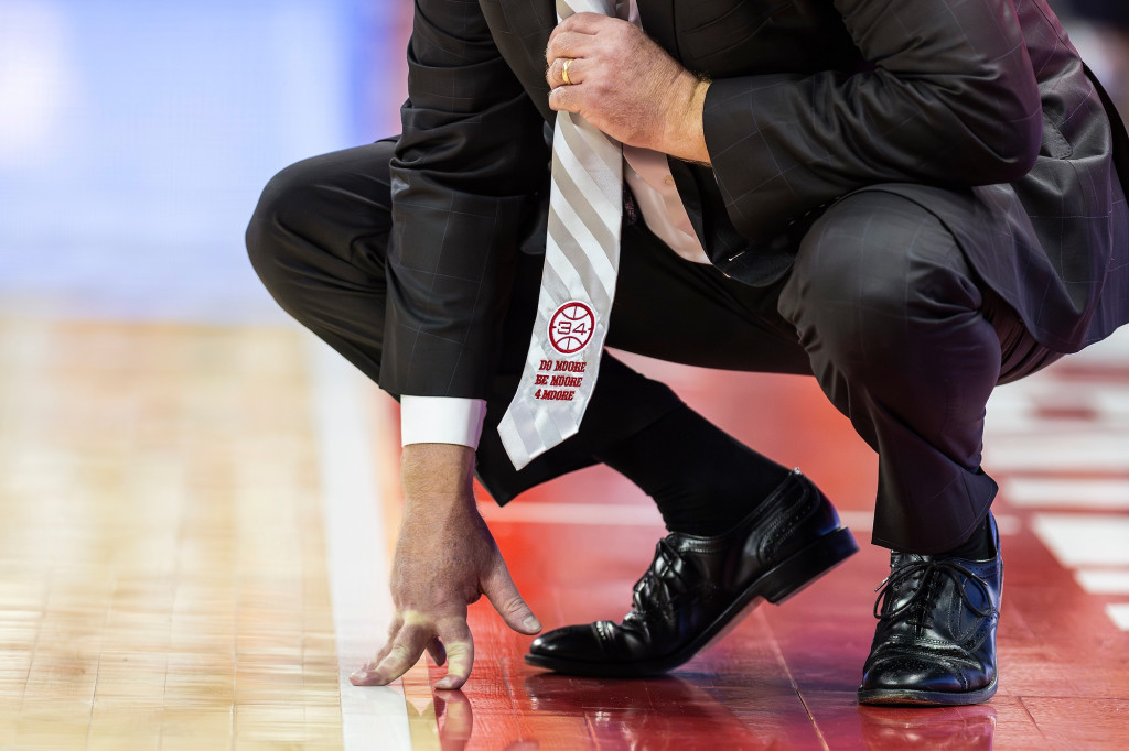 A coach wearing a suit and tie crouches on the sidelines.