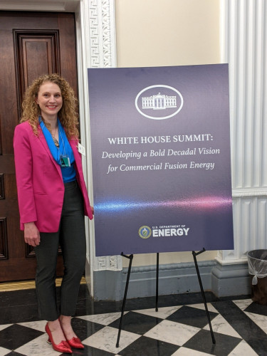Photo of Steffi Diem in a pink blazer and black pants smiles as she stands next to a sign that reads White House Summit: Developing a Bold Decadal Vision for Commercial Fusion Energy. She is standing in a corridor of the White House.