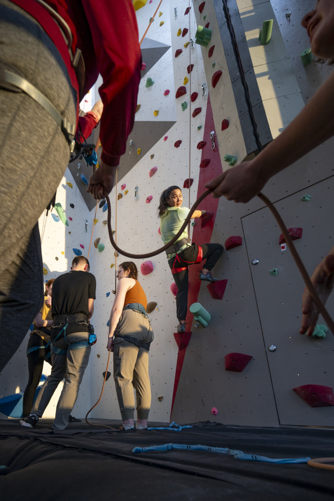 A person climbs up a climbing wall, with ropes attached.