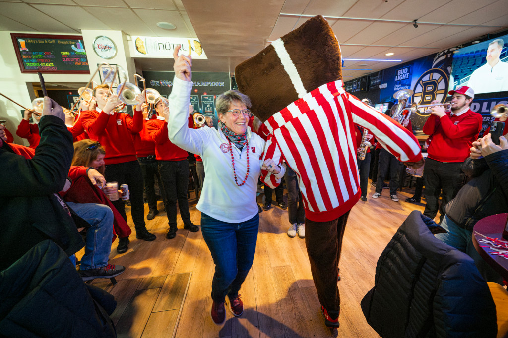 A woman wearing a Wisconsin shirt links arms with Bucky Badger and dances.