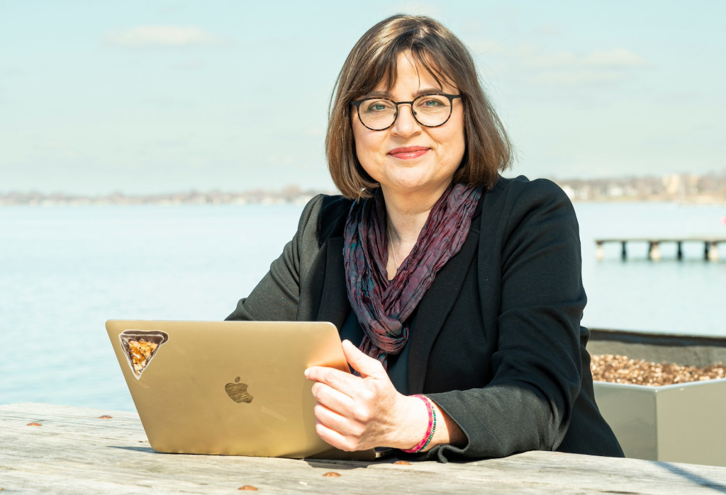 A woman sits at a desk with a laptop; behind her is Lake Mendota.