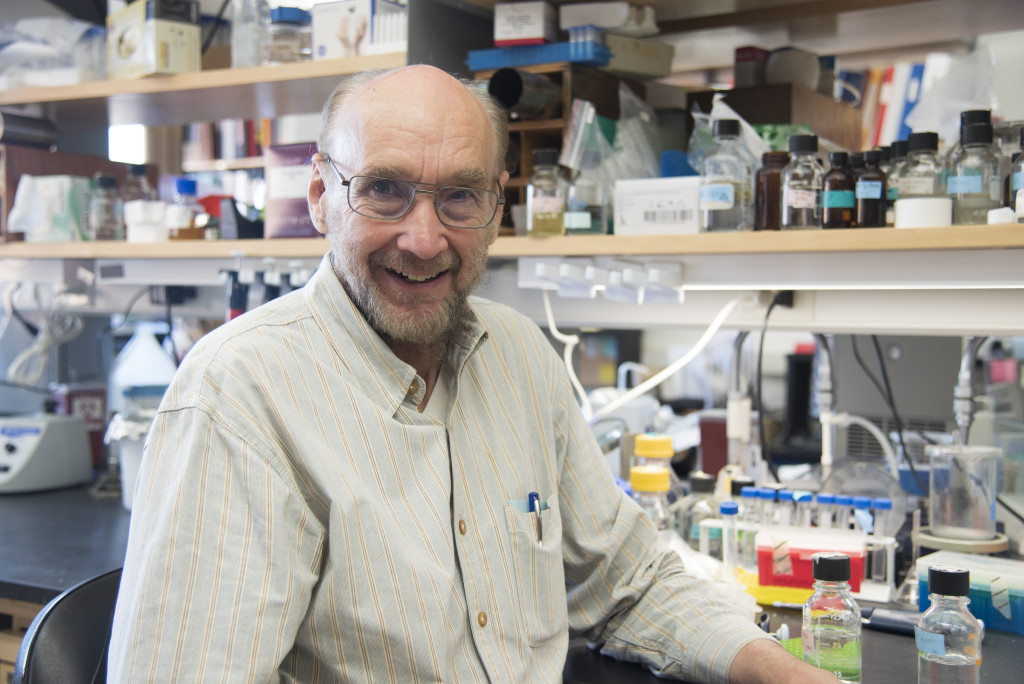 A man smiles at the camera, behind him are shelves full of lab equipment.