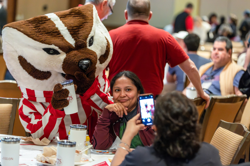 A woman sitting at a table poses with Bucky Badger.