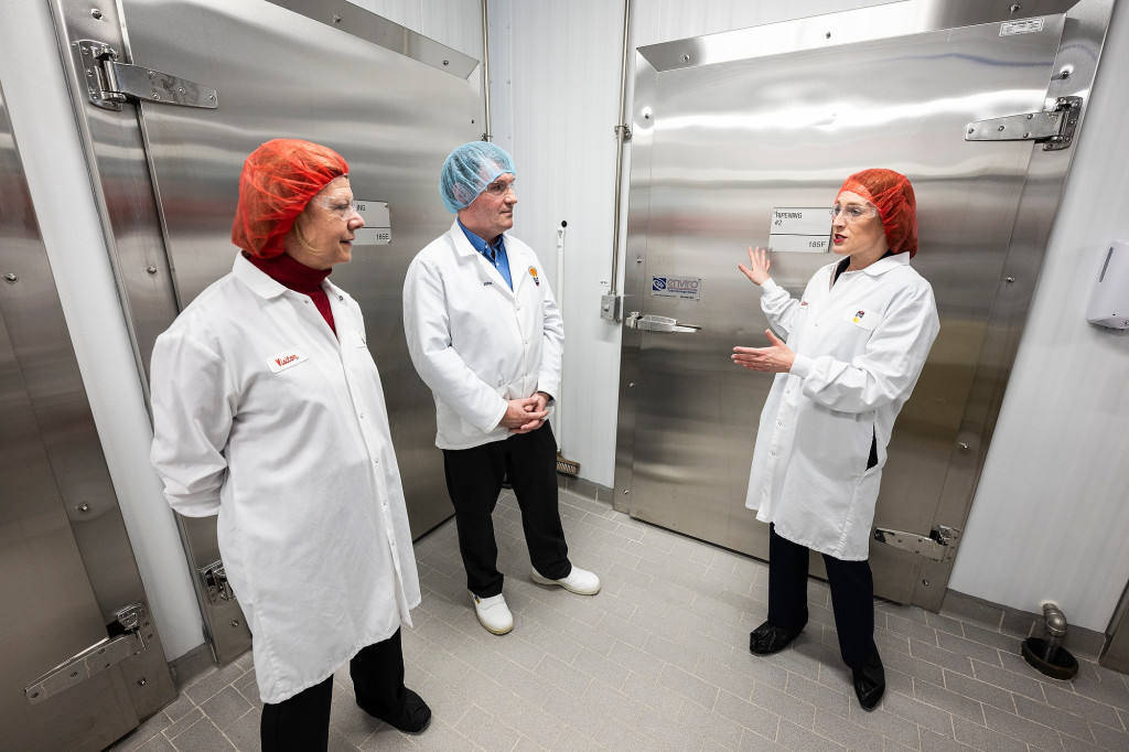 Three people wearing white coats and hair nets talk.