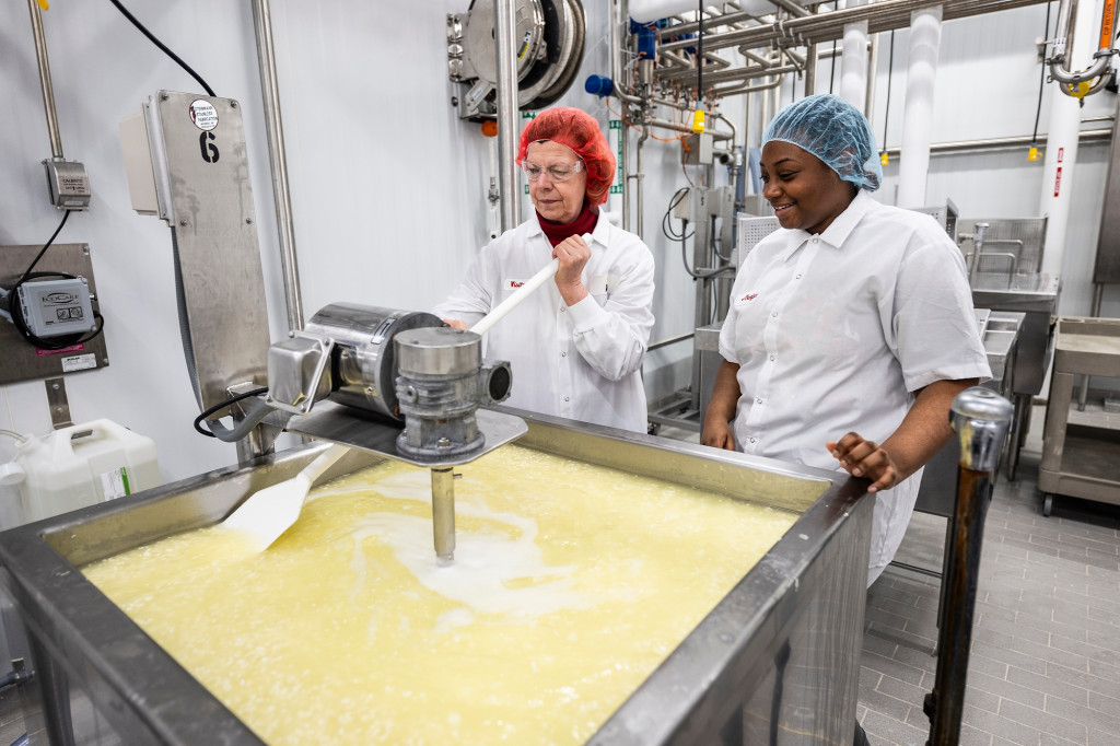 Two women stand by a vat of yellow liquid, and one stirs it with a paddle.