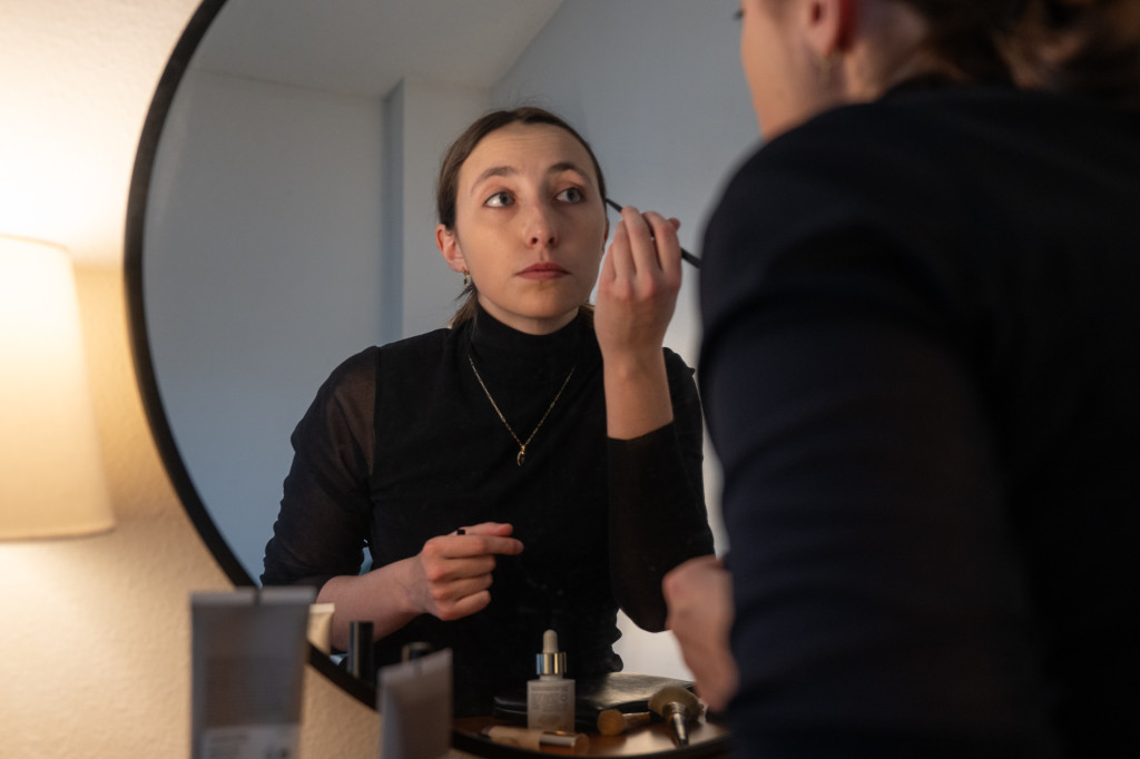 A woman applies makeup to her face as she peers in the mirror.
