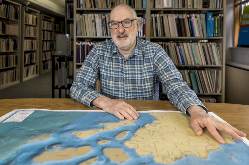 David Hart sits at a table in the library stacks with his hands on a topographical map
