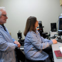 A man and a woman in blue lab coats look at an image on a screen in a laboratory.