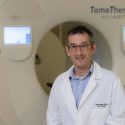 A photo of Jonathan Levine wearing a white lab coat and standing in front of a CT scanner.