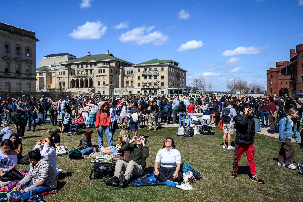 Dozens of people relax on a grassy field, many looking up at the sky.