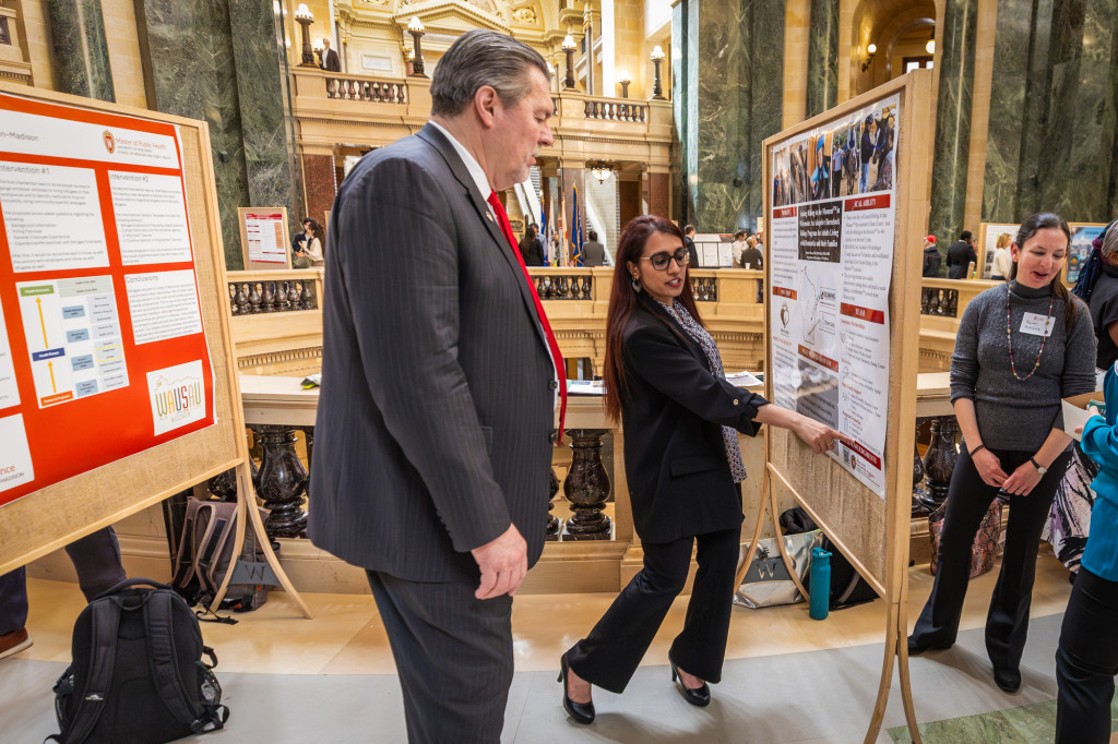 A woman gestures to a poster board and talks to a man in a suit.