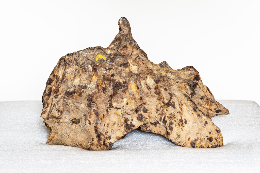 The meteor on display against a white background. It resembles a small mountain peak in shades of tan, brown and rust.