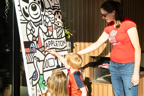 A woman watches as two children color in a large black and white poster of Bucky Badger and a flamingo surrounding the word Appleton.