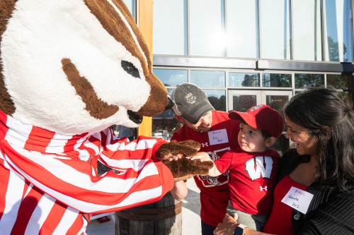 Two UW alumni dressed in red shirts hold their young son up to shake hands with Bucky Badger.