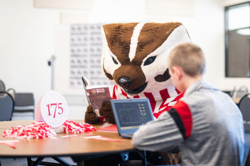 Bucky Badger sits at a table and reads while a seventh-grade student works on a laptop.