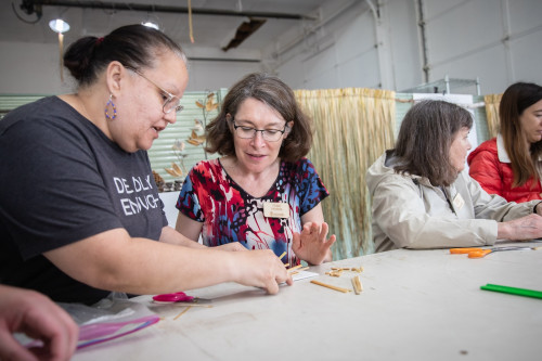 Two women sit at a table and work together on a weaving project.