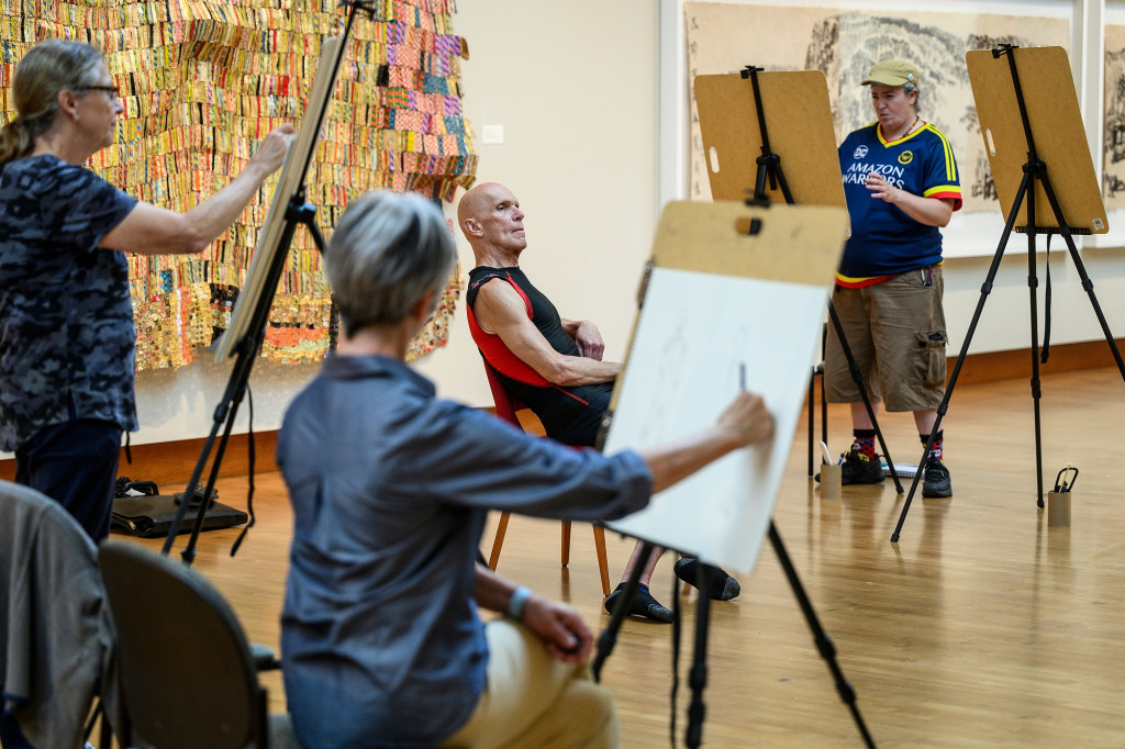 Three people sketch at easels as a model poses.