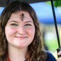 A woman smiles to the camera as a periodical cicada rests on her forehead.