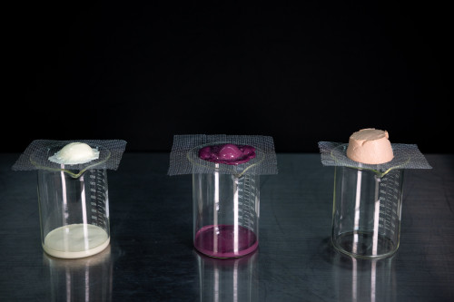 A photo of an experimental setup to test the melting speed of ice cream. Three glass beakers topped with a piece of wire mesh each support a scoop of ice cream. The ice cream melts through the mesh and into the beakers.