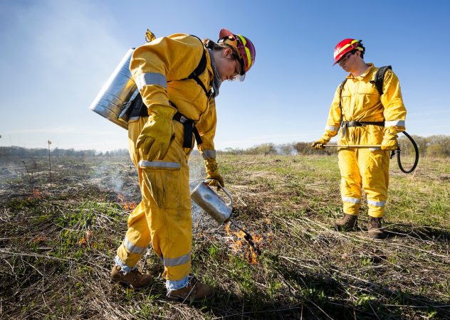 Two people in yellow firefighter outfits light a prairie field on fire.