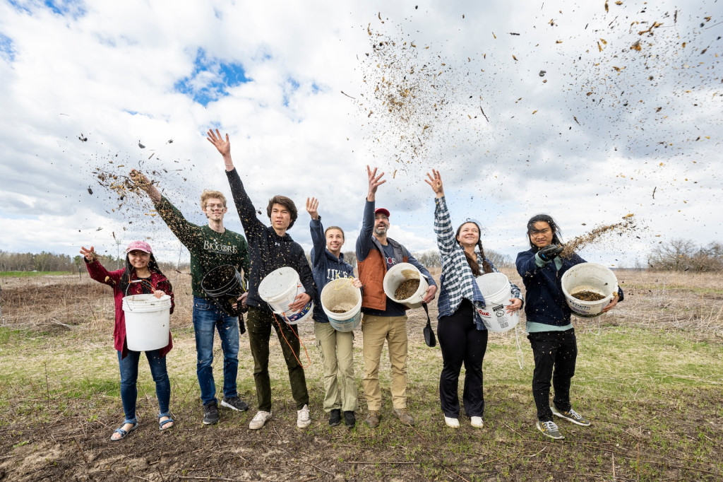 Seven students stand in a line in a field and throw seeds into the air.