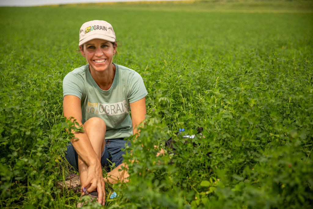 A woman crouches in a field full of green vegetation and smiles at the camera.