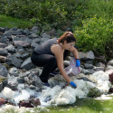 A woman crouches at the edge of a lake, collecting foam that's gathered there.