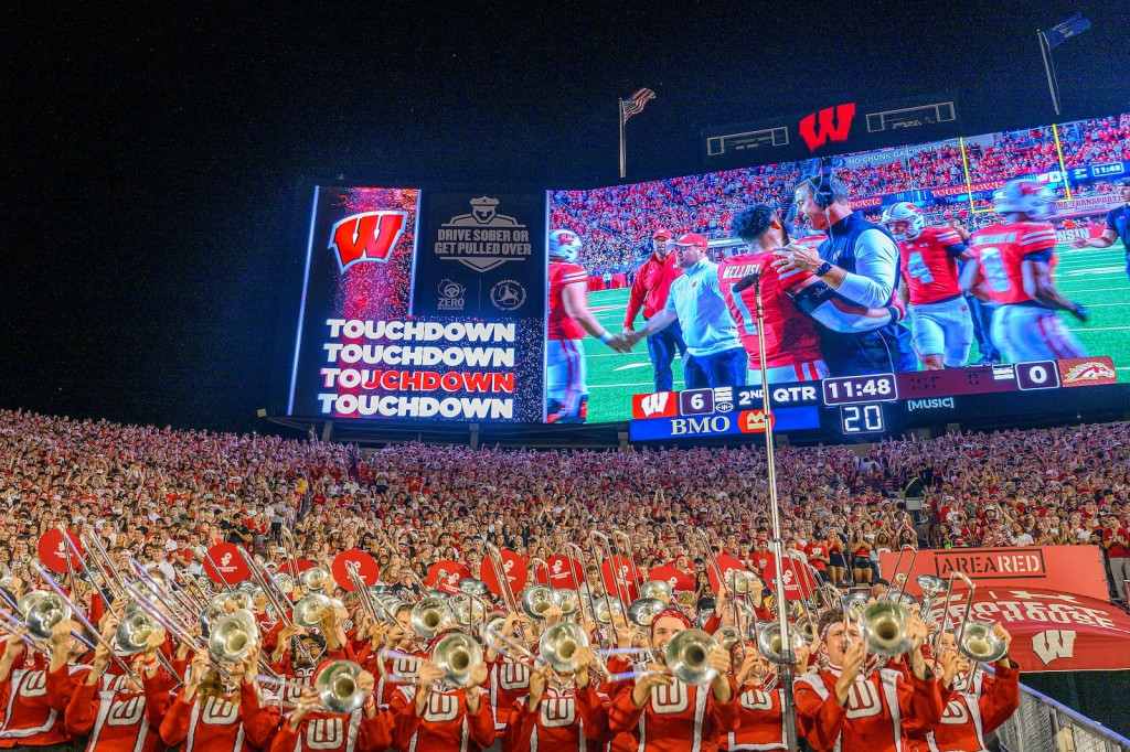 People wearing red marching bad uniforms play music in a stadium; a scoreboard in the background replays a touchdown play.