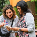 Two women huddle over a cell phone, sharing contacts.