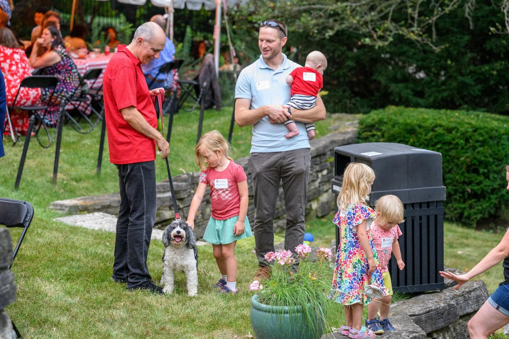 Two men speak, one holds a child. Three children and a dog stand around at their feet.