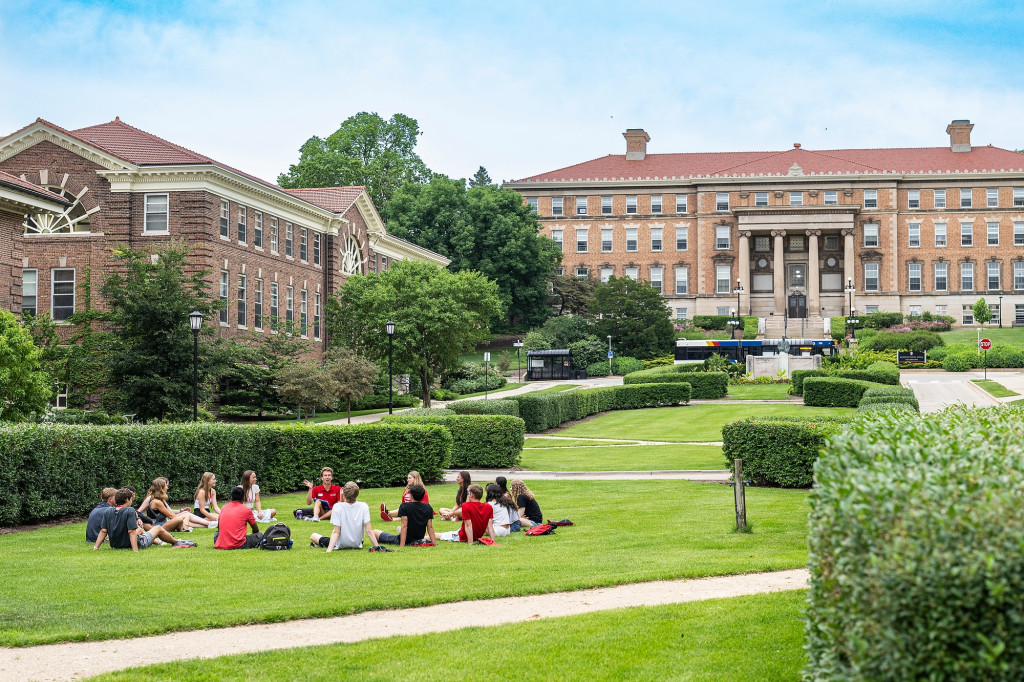 A far away view of a group of college students sitting in a circle on a grassy green hill surrounded by older academic buildings.