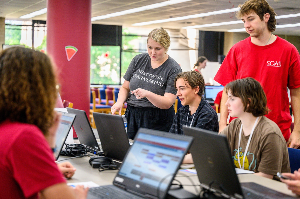 A standing woman leans over two seated college students and points at their computers.