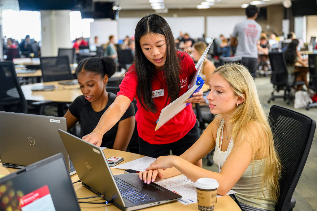 A standing woman leans over two seated college students and points at their computers.