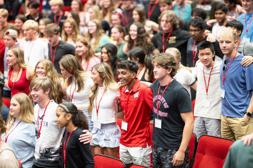 A room full of college students stand together in a line with arms linked around each other while singing a song.