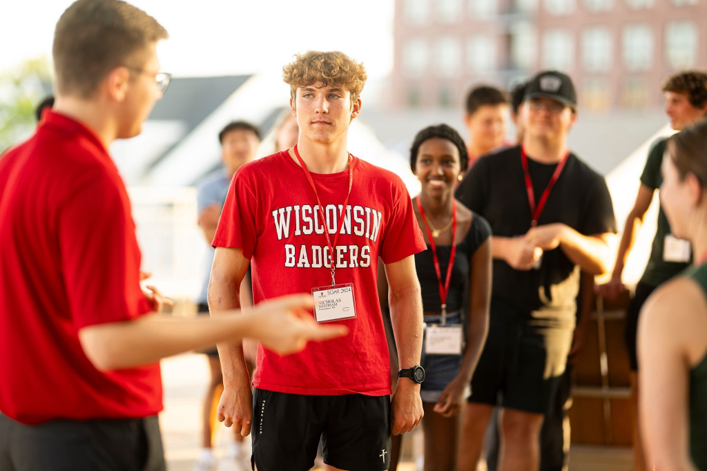 A student in a red UW Badgers shirt listens along with fellow incoming students as they listen to instructions for an activity.