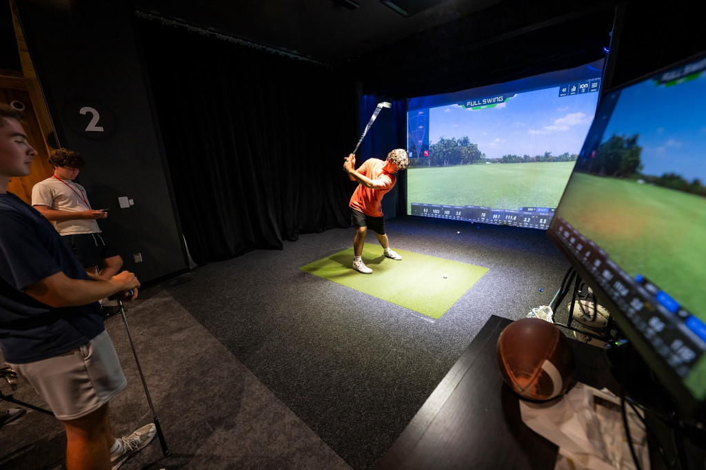 A man stands in the center of a dark room holing a golf club while playing a virtual golf game.