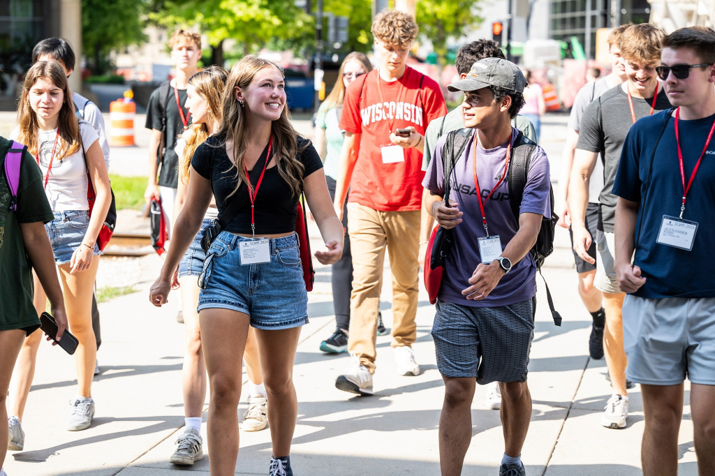 Two college students wear red lanyards happily talk to each other while crossing the street.