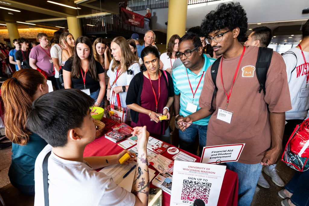 A group of people stand one one side of a table while conversing with two people sitting on the opposite side of the red-covered table.