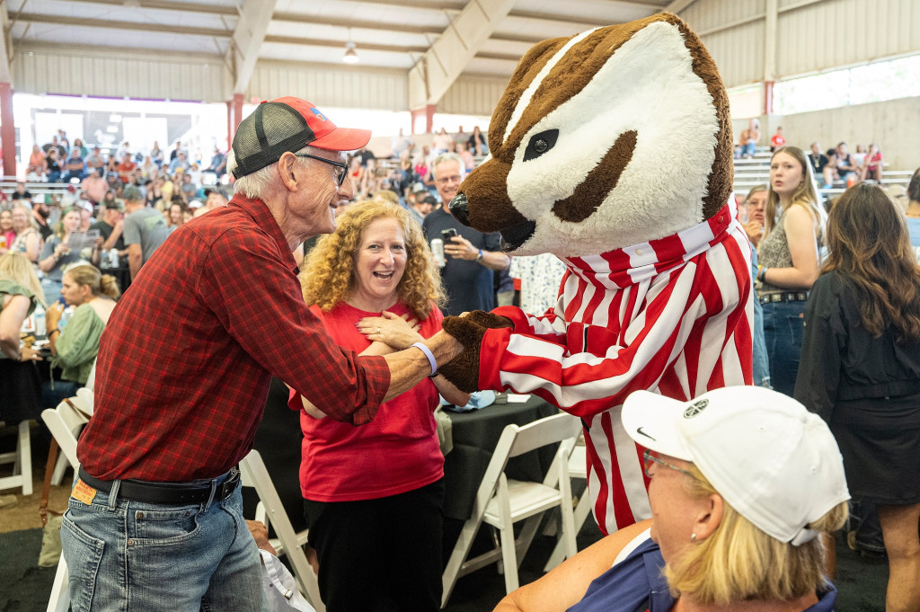 A man reaches out for a handshake from a Bucky Badger mascot. A woman stands behind them laughing.