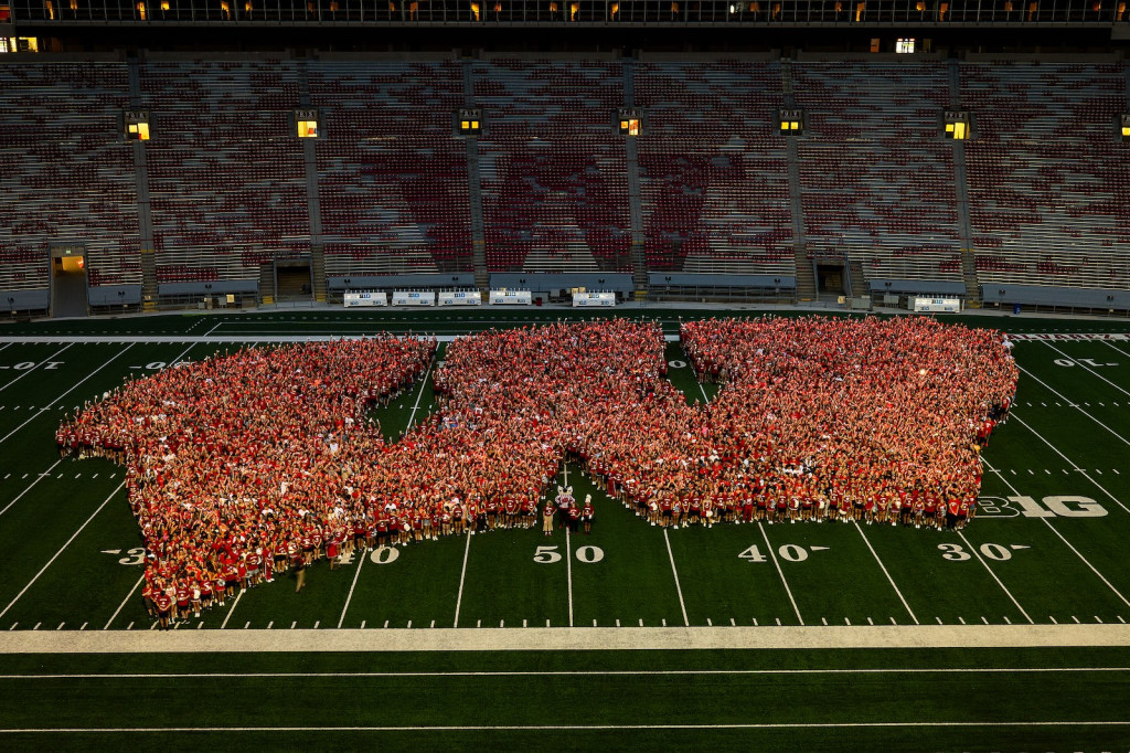 A large group of students wearing red and white clothes form a W on a football field.