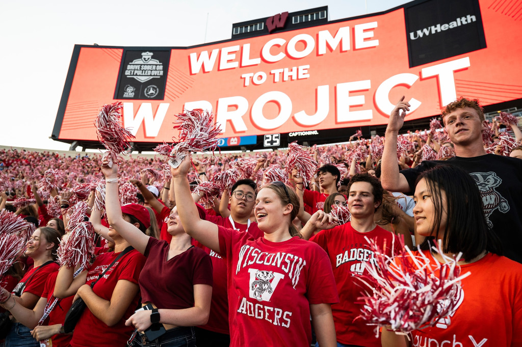 People wearing Wisconsin Badger shirts cheer and laugh as they stand in a stadium.